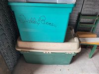 Photo showing two large plastic storage totes stacked, one teal tote labeled 'Teddy Bears' with a teal lid on top of a two-tone green and beige tote with a beige lid.