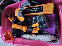 View into pink plastic bin holding multiple Halloween decorations including a black and orange plush spider, white tinsel garland, and boxed Halloween string lights and decorations.