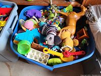 Blue bin filled with a variety of children's toys including plush animals, action figures, and plastic toy tools.