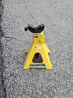 Yellow metal car jack stand on asphalt surface with black adjustable cradle at the top.