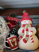 Photo showing large ceramic snowman cookie jar wearing a red and green scarf and hat, holding a candy cane, along with ceramic Santa Claus mug
