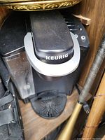 Black Keurig coffee maker placed on wooden shelf, showing top brewing lid with Keurig logo, water reservoir, and drip tray.