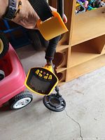 Metal detector held by a hand, showing yellow control box with LCD screen, buttons, black circular search coil, and adjustable strap.