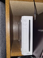 Front view of the white Nintendo Wii console showing buttons and disc slot, placed in a wooden shelf next to a Samsung device.
