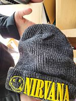 Photo showing the black ribbed knit beanie hat held up, clearly showing bright yellow Nirvana embroidered text and smiley face logo.