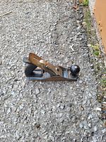 Photo of a metal hand plainer tool placed on asphalt ground showing front knob and wooden rear handle.