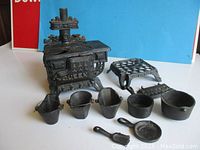 Front angle photo showing the cast iron miniature stove with the front embossed word 'Queen,' two pots, one pan, three coal buckets, and small cast iron shovel.