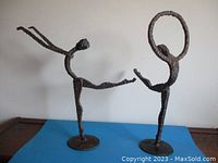 Front view of two welded steel dancer statues showing abstract elongated limbs and textured finish on a blue surface.