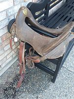 Side view of dark brown leather McClellan saddle hanging on black bench. Shows saddle seat, leather straps, and hardware.
