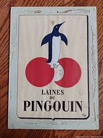 Frontal photo of antique Laines du Pingouin print featuring stylized penguin standing on red balls of yarn. Print signed by Will Lacroix, under glass with light blue distressed wooden frame.