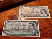 Photo showing two Canadian one dollar bills placed on a wooden surface; top bill is the 1973 last issue, bottom bill is the 1967 Centennial issue.