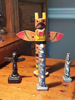 Front view of one colorful hand-painted wooden totem pole and three smaller pewter/cultured argillite totem pole sculptures on wooden surface.