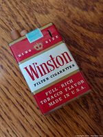 Front and angled views of the replica Winston filter cigarette pack lighter showing the red and white design with gold trim and tax stamp detail.