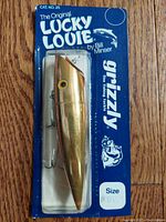 Single gold Lucky Louie lure in packaging against wood floor