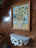 Wall shelf with ornate white scrollwork and slightly curved top seen resting on wooden floor with framed floral print nearby.