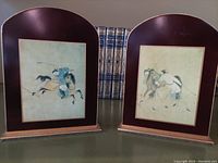 Front view of both bookends showing Mongol polo horsemen illustrations with a burgundy background and wooden base, placed side by side with decorative books behind.