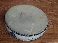 Side view of drum showing circular painted fairy/bird design on worn natural hide stretched over wooden frame with metal button rim on hardwood floor
