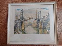 Framed artwork depicting a bridge over water with boats and houses, shown angled on wooden floor.