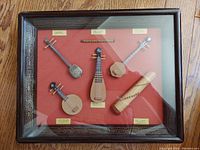Frontal view of the shadow box showing all six miniature handcrafted Chinese musical instrument replicas with labels on a red background inside a wooden frame.