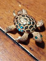 View of golden sea turtle brooch showing textured body and emerald green eyes, placed on wooden surface