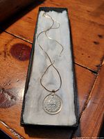 Full view of the coin pendant on the braided sterling silver chain displayed in a black lined box on a wooden surface.