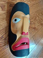 Left angled view of the handcrafted wooden warrior mask showing painted face details and black painted hair/head covering.