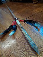 Full view of the lance showing wooden shaft, steel blade tip and feather decorations on hardwood floor.
