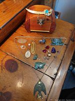 Top view of the cedar treasure chest with multiple small jewelry items and collectibles laid in front. Items include necklaces, pins, small figurines, and decorative stones.