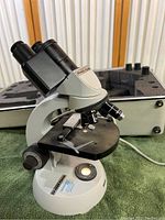 Angled view of Carl Zeiss binocular microscope with objectives and stage, set on green cloth, with hard case behind.