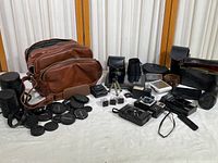 Wide view of all included items laid out on a white surface: brown leather bag, multiple lens caps, pouch cases, tripod adapters, a strobe flash, several small electronic parts, and broken cameras.