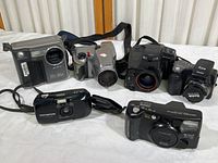 Six cameras arranged together on a surface showing front and top views including Sony Mavica, Olympus D-500L, Kodak Easyshare, Fuji 1000 Zoom, Olympus Stylus, and ZoomCam camera.