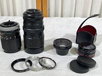 Wide view of all lenses and adapter rings laid out showing various brands and sizes. Includes Canon FD 135mm lens, Bushnell zoom lens, Sony wide lens, Vivitar video lens in leather case and metal rings.