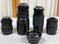Five camera lenses displayed upright and lying on a surface, showing size and styling differences.