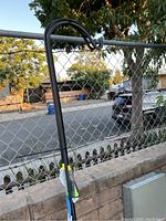 Photo of a black metal shepherds hook in front of a chain link fence and stone wall, showing curved top hooked for hanging and middle section of the pole.