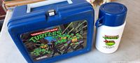 Front view of TMNT blue plastic lunch box with ninja turtle graphic and matching white and blue thermos