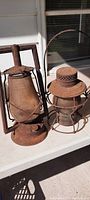 Two primitive antique railroad lanterns on a white surface, showing their rusted exteriors, metal handles, and intact glass globes.