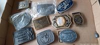 Overview of the belt buckle lot showing twelve vintage metal belt buckles of about 4 inches, some wrapped in plastic packaging