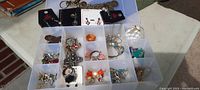 Photo of sectioned plastic jewelry case showing various compartments filled with mixed costume jewelry including bracelets, earrings, necklaces, and brooches.