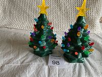 Pair of green ceramic Christmas trees about 8 inches tall with multicolored bulbs and yellow star on top, placed against a neutral woven background.