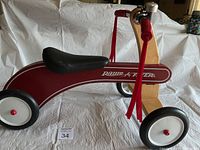 Front and side view of vintage style wooden Radio Flyer balance bike with red body, black seat, black wheels with white rims, red tassels on handlebars, and a silver bell.