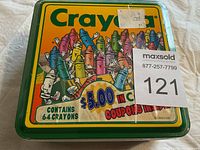 Sealed green metal Crayola tin with cartoon crayon characters, marked as containing 64 crayons and featuring a $5.00 coupon offer.