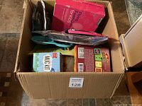 View showing the contents of a cardboard box filled with boxed and packaged items including kitchenware and craft kit