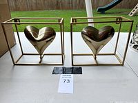 Two gold-tone metal heart figurines each in an open rectangular metal frame sit on a white table outdoors.