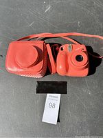 Coral pink Instax Mini 7+ camera next to matching leather carrying case with shoulder strap on a dark surface