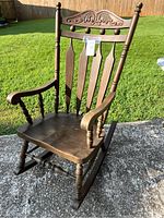 Front view of large solid wood rocking chair showing carved top rail, turned spindles, and curved armrests.