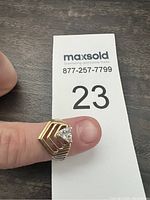 Ring placed on finger with maxsold card showing lot number 23, displaying front view of the ring with stone and stepped design.