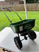 Side-left angle showing the full spreader with two wheels, the green hopper with Scotts branding, and metal handlebar.