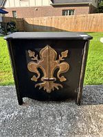 Front view of large black wooden chest with prominent gold fleur-de-lis decoration on doors.