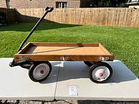 Side view of vintage wooden wagon showing rectangular bed, metal handle, and four rubber tires on white rims.