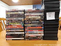 Photo showing a large stack of DVDs arranged on a table with titles visible including Schindler's List, Gladiator, Lethal Weapon, The Godfather, and others. Also shows a separate stack of about 30 empty black DVD cases.